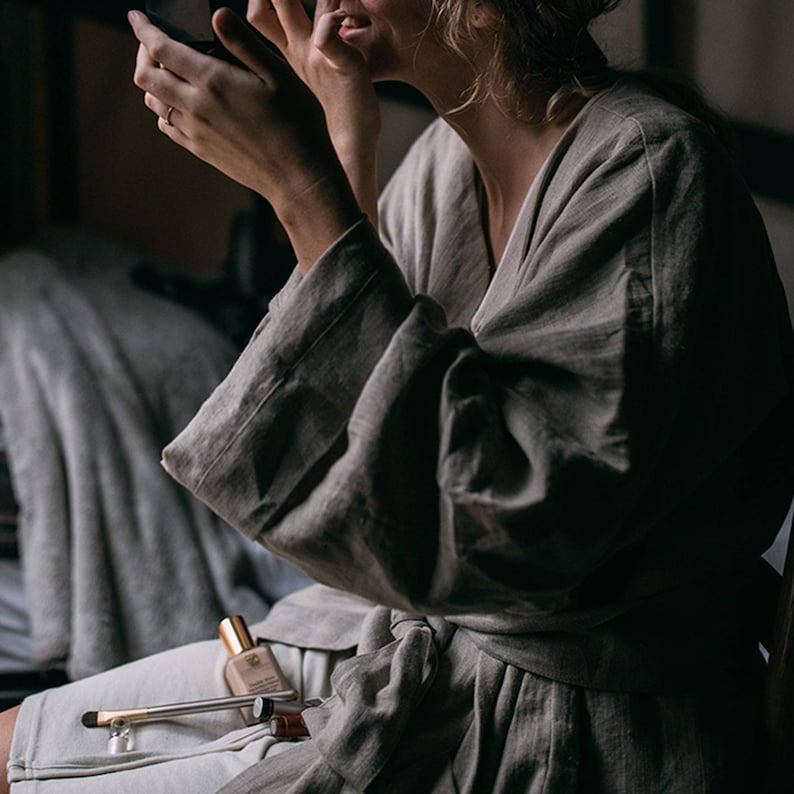 May include: A person wearing a light brown linen robe is applying makeup while sitting on a bed.  A small bottle of foundation, a makeup brush, and a lipstick are on the bed.
