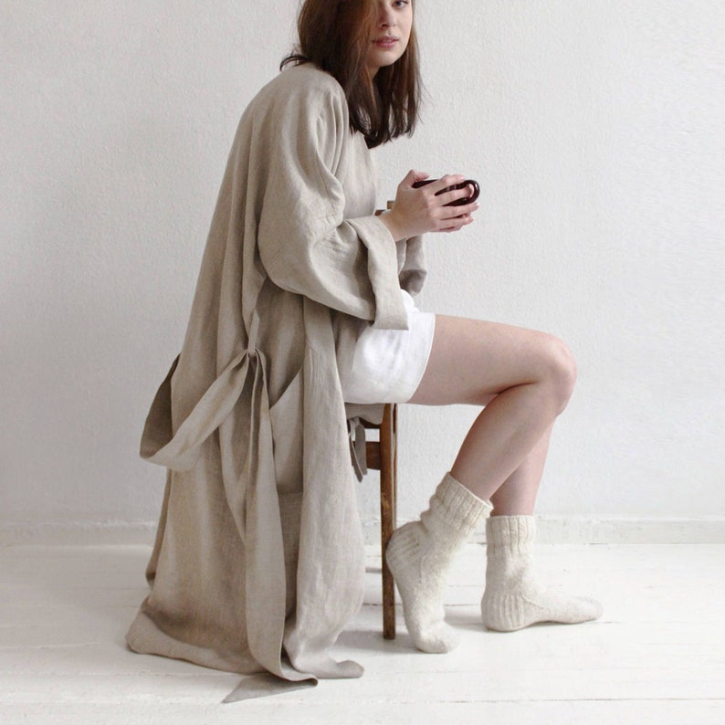May include: A woman wearing a beige linen robe with a white belt and white socks. She is sitting on a wooden stool and holding a cup.