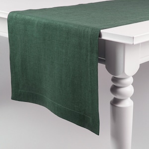 May include: A dark green linen table runner with a subtle texture. The runner is folded and draped over a white table with a turned leg.