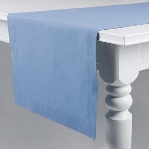 May include: A light blue linen table runner with a white border. The runner is laying on a white table with a turned leg.