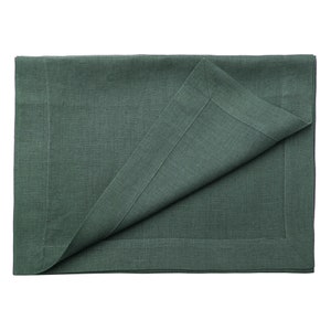 May include: A rectangular dark forest green linen placemat featuring a visible woven texture and a mitered corner border detail. One corner of the placemat is folded over to reveal the underside, showcasing the consistent solid green color.