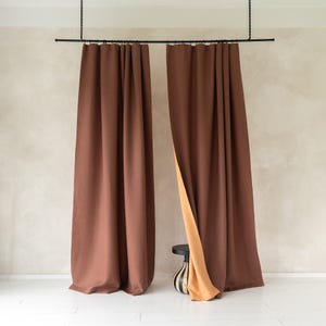 Puede incluir: Dos cortinas marrones, largas hasta el suelo, colgadas de una barra negra. Una cortina está parcialmente abierta, revelando un forro beige. Las cortinas están en una habitación con una pared de color neutro y suelo blanco. Se ve una pequeña mesa negra con un jarrón a rayas.