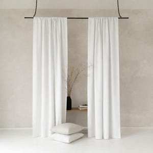 Op de afbeelding: Twee witte, vloerlange gordijnen hangen aan een zwarte stang die aan kettingen is bevestigd. Een zwarte vaas met gedroogde planten staat op een houten plank, met twee vierkante kussens in neutrale kleuren op de vloer. De achtergrond is neutraal beige.