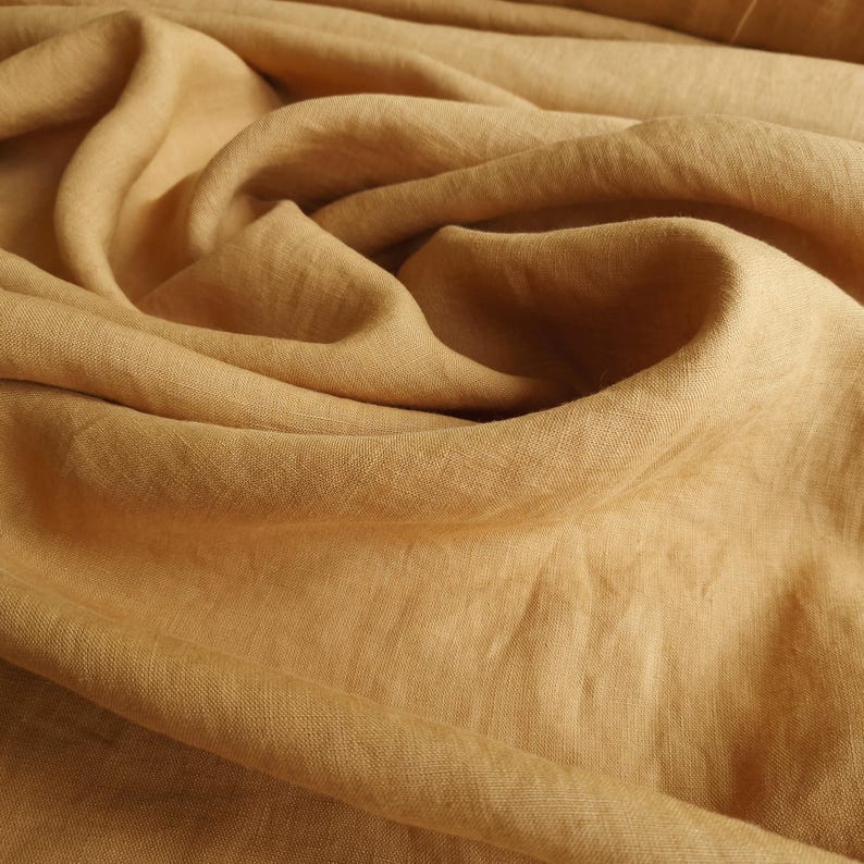 May include: Close-up of a tan-colored linen fabric with soft folds and a textured surface. The fabric is a warm, neutral tone, and the folds create shadows and highlights, adding depth to the image. The material appears to be lightweight and breathable.