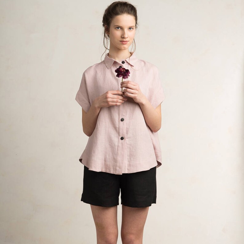 May include: A pink linen shirt with a button-down front and short sleeves. The shirt has a relaxed fit and is slightly oversized. The model is holding a single dark purple flower.