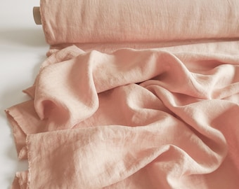 May include: A roll of blush pink linen fabric, partially unrolled to reveal the textured material. The fabric has a soft, flowing drape with visible wrinkles and a raw edge. The color is a muted, light pink.