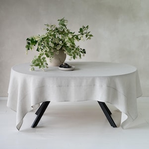 May include: A round, light gray linen tablecloth draped over a table with black legs. A vase with white flowers and greenery sits on the table, along with a small plate of dark berries. The table is set on a white floor.