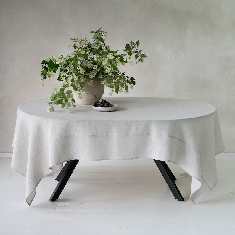 May include: A round, light gray linen tablecloth draped over a table with black legs. A vase with white flowers and greenery sits on the table, along with a small plate of dark berries. The table is set on a white floor.