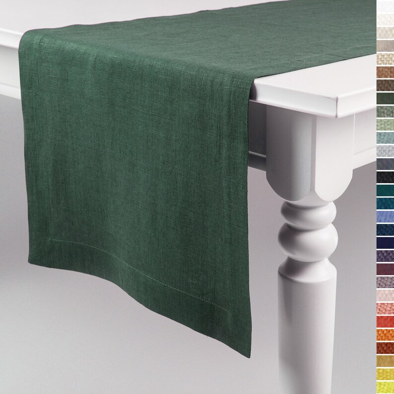 May include: A dark forest green table runner with a visible woven texture and a simple hemmed border. The runner is draped over the edge of a white table, showcasing the fabric's weave and the deep, solid green color of the textile material.