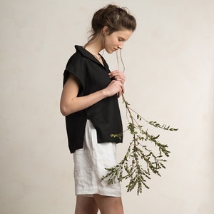 Peut inclure: Une femme portant un haut en lin noir avec un col et un short en lin blanc. Elle tient une branche avec des feuilles vertes.