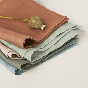 Pode incluir: Um conjunto de quatro guardanapos de linho em tons de marrom, verde e azul. O guardanapo superior está dobrado e tem uma cápsula de papoula seca no topo.