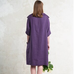 May include: A purple linen dress with a collared neckline and short sleeves. The dress has a relaxed fit and falls to the knees.