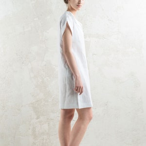 May include: A woman wearing a white linen dress with short sleeves. She is standing in front of a white textured wall.