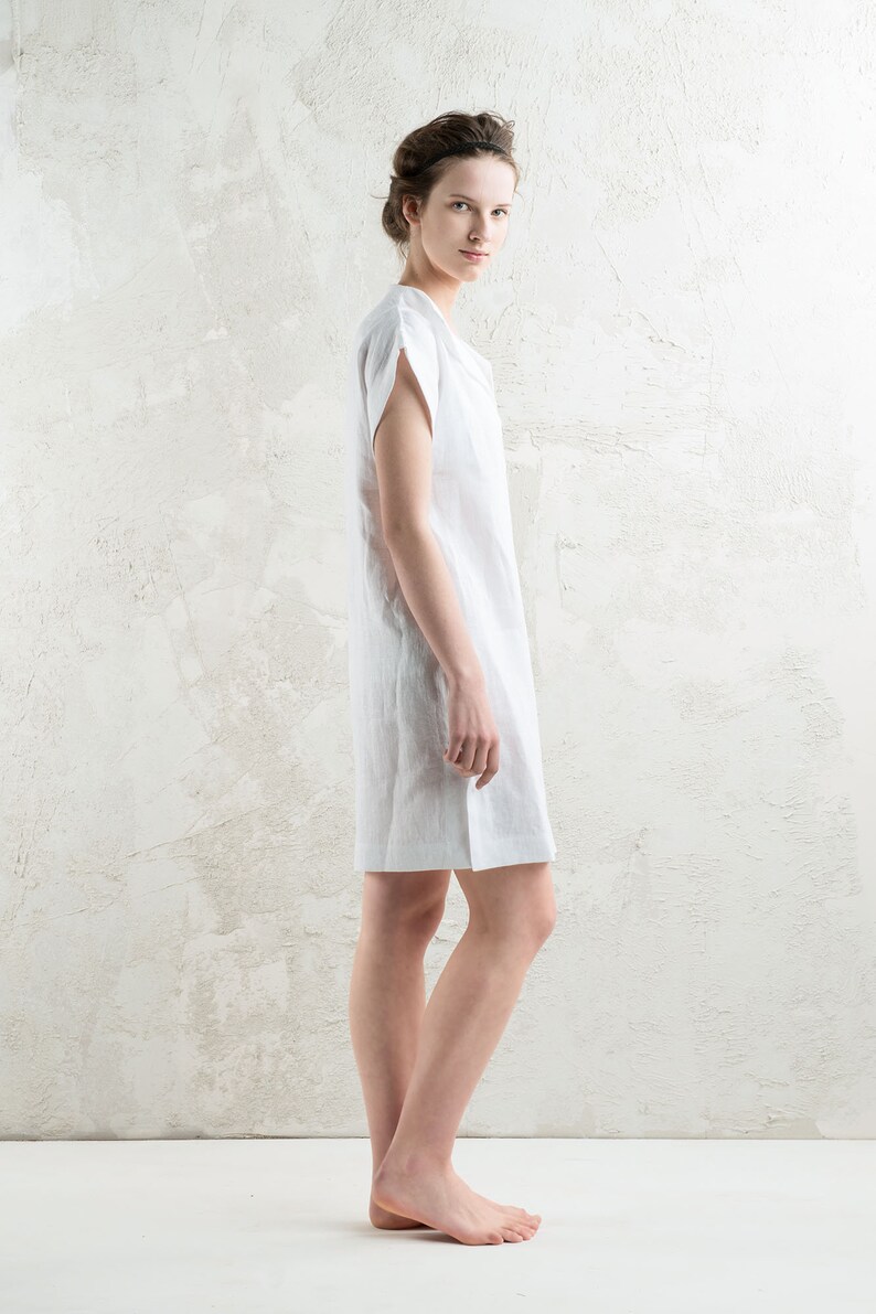 May include: A woman wearing a white linen dress with short sleeves. She is standing in front of a white textured wall.