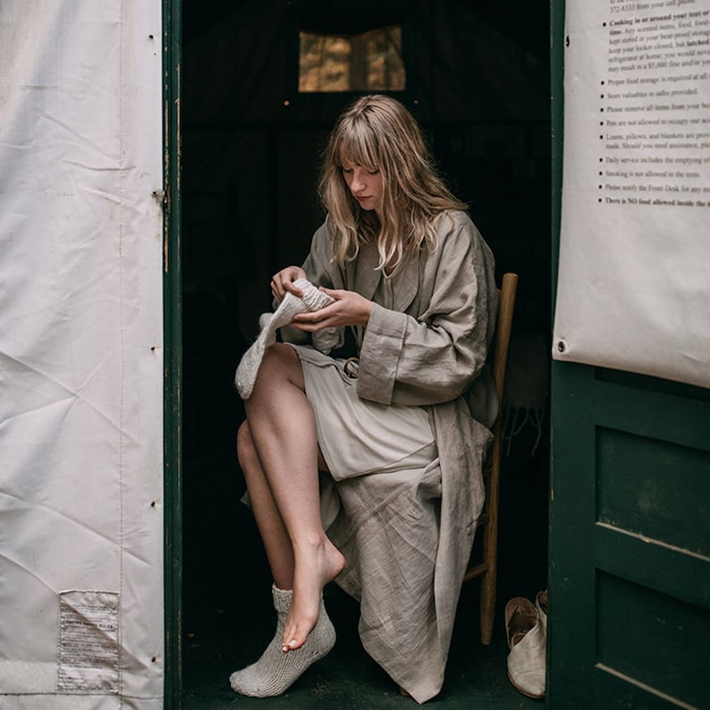 May include: A woman sits in a chair inside a tent, wearing a light brown robe and white socks. She is holding a white knitted item in her hands.