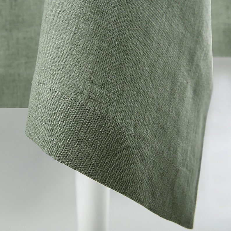 May include: Close-up of a sage green linen tablecloth. The fabric has a textured appearance with visible weave details. The edge of the tablecloth is neatly hemmed, and the corner is visible, suggesting a square or rectangular shape. The tablecloth is draped over a white surface.