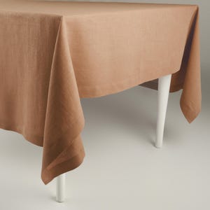 May include: A rectangular, light brown tablecloth draped over a white-legged table. The fabric has a textured appearance, with folds and shadows adding depth. The tablecloth covers the table, with the edges hanging down.