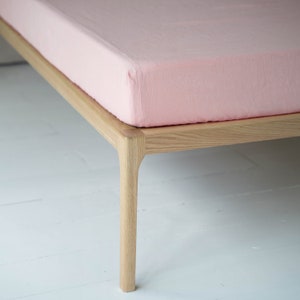 Puede incluir: Primer plano de una sábana de cama de lino rosa claro sobre un marco de cama de madera. El marco de la cama tiene un diseño simple y moderno con bordes redondeados.