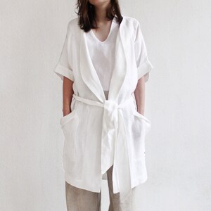 May include: A white linen robe with a tie belt and two pockets. The robe has a relaxed fit and a long, flowing silhouette.