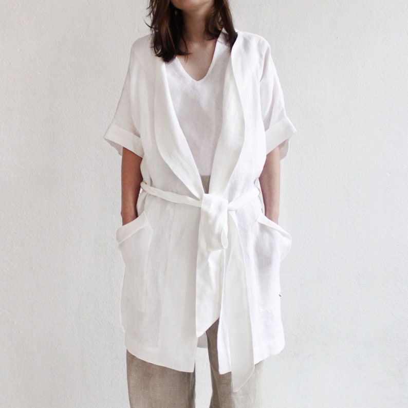 May include: A white linen robe with a tie belt and two pockets. The robe has a relaxed fit and a long, flowing silhouette.
