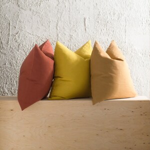 May include: Three square throw pillows in shades of terracotta, yellow, and tan. The pillows are arranged on a light wood surface against a textured white wall. The pillows have a soft, inviting appearance.