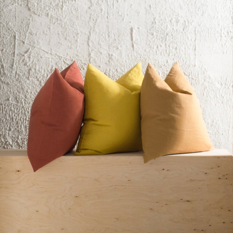 May include: Three square throw pillows in shades of terracotta, yellow, and tan. The pillows are arranged on a light wood surface against a textured white wall. The pillows have a soft, inviting appearance.