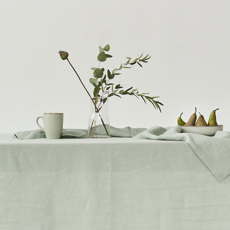 Peut inclure: Une nature morte paisible avec une nappe en lin vert clair. Un vase en verre transparent contient des branches d'eucalyptus. Une tasse en c&eacute;ramique et un bol de poires compl&egrave;tent la sc&egrave;ne. L'ensemble &eacute;voque le calme et la simplicit&eacute;, id&eacute;al pour la d&eacute;coration int&eacute;rieure.