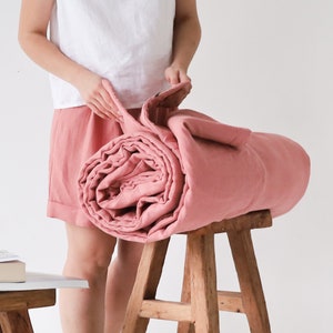 May include: A rolled-up, dusty rose-colored blanket rests on a wooden stool. The blanket is made of a soft, textured fabric. The person holding the blanket is wearing a white top and pink shorts. The stool is made of light-colored wood.