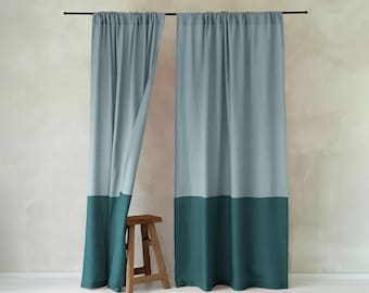 May include: Two floor-length curtains hanging from a black rod, featuring a color-blocked design with a light blue-gray upper section and a solid dark teal lower section. A small, four-legged wooden stool sits in front of the left curtain.