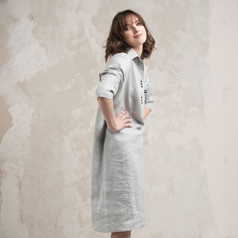 May include: A woman wearing a light grey linen dress with a button-down front and a slightly loose fit.