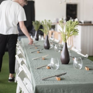 Puede incluir: Una mesa decorada con un mantel verde salvia, con copas de vino sin pie, cuchillos plateados y pequeños reposacuchillos de madera. Jarrones marrones oscuros contienen ramitas verdes, creando una estética elegante y minimalista. Sillas plegables blancas.
