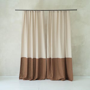 Puede incluir: Cortinas de dos paneles en beige y marrón colgadas de una barra negra. La parte superior de las cortinas es de color beige liso, mientras que el tercio inferior es de color marrón liso. Las cortinas cuelgan frente a una pared de color beige claro. Miden unos 200 cm.