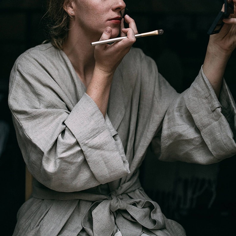 May include: A person wearing a light gray linen robe with a sash tied at the waist. They are applying makeup with a brush and looking in a mirror.