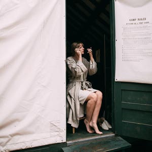 May include: A woman in a light gray robe sits in a wooden cabin doorway, applying makeup while looking in a small mirror. The cabin door is green and has a sign that reads "Camp Rules: Staying in a Tent Cabin".