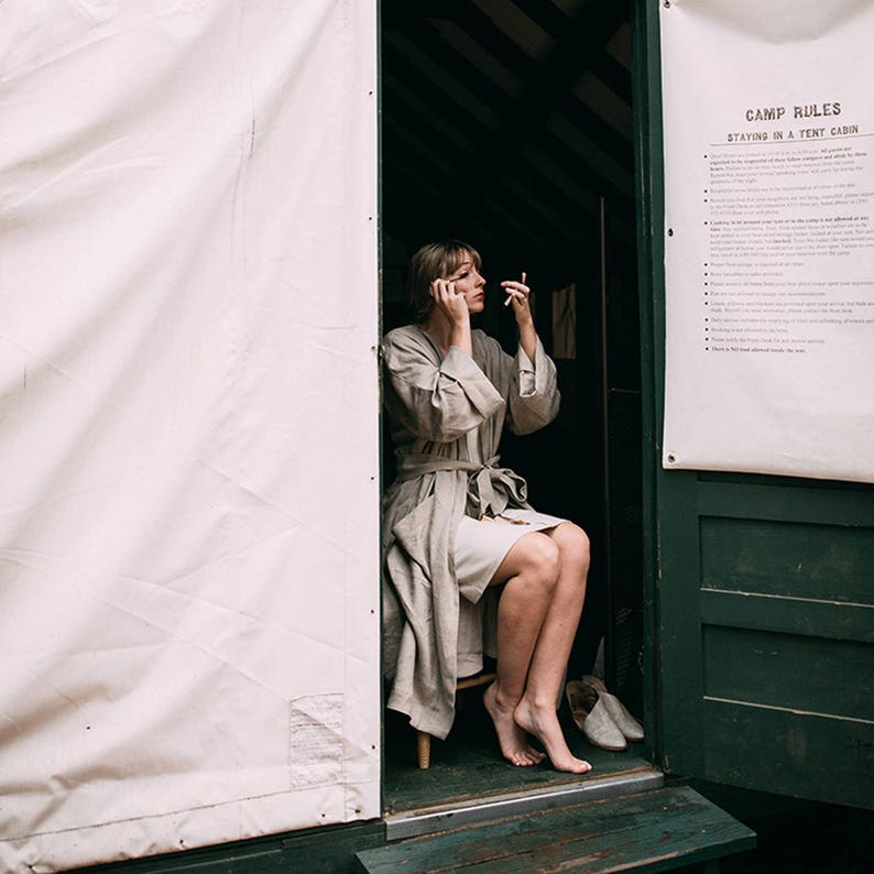May include: A woman in a light gray robe sits in a wooden cabin doorway, applying makeup while looking in a small mirror. The cabin door is green and has a sign that reads "Camp Rules: Staying in a Tent Cabin".