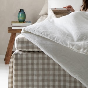 May include: A white bed with a gingham bed skirt in a beige and white pattern. The bed is made with white linen bedding and a book is resting on top of the bedding.