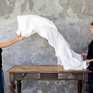 May include: Two people stand on either side of a wooden table. One person is holding a white linen sheet in the air. The sheet is flowing and billowing. The other person is standing with their hands on their hips. The background is a gray concrete wall.