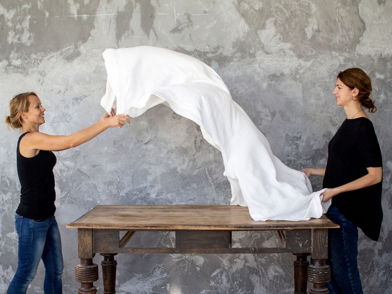 May include: Two people stand on either side of a wooden table. One person is holding a white linen sheet in the air. The sheet is flowing and billowing. The other person is standing with their hands on their hips. The background is a gray concrete wall.