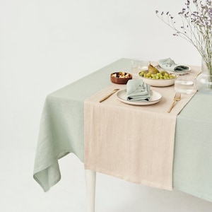 May include: A dining table set for a meal, featuring a light green tablecloth with a beige runner. The table is set with plates, napkins, glasses, and a bowl of fruit. A vase of purple flowers adds a decorative touch.