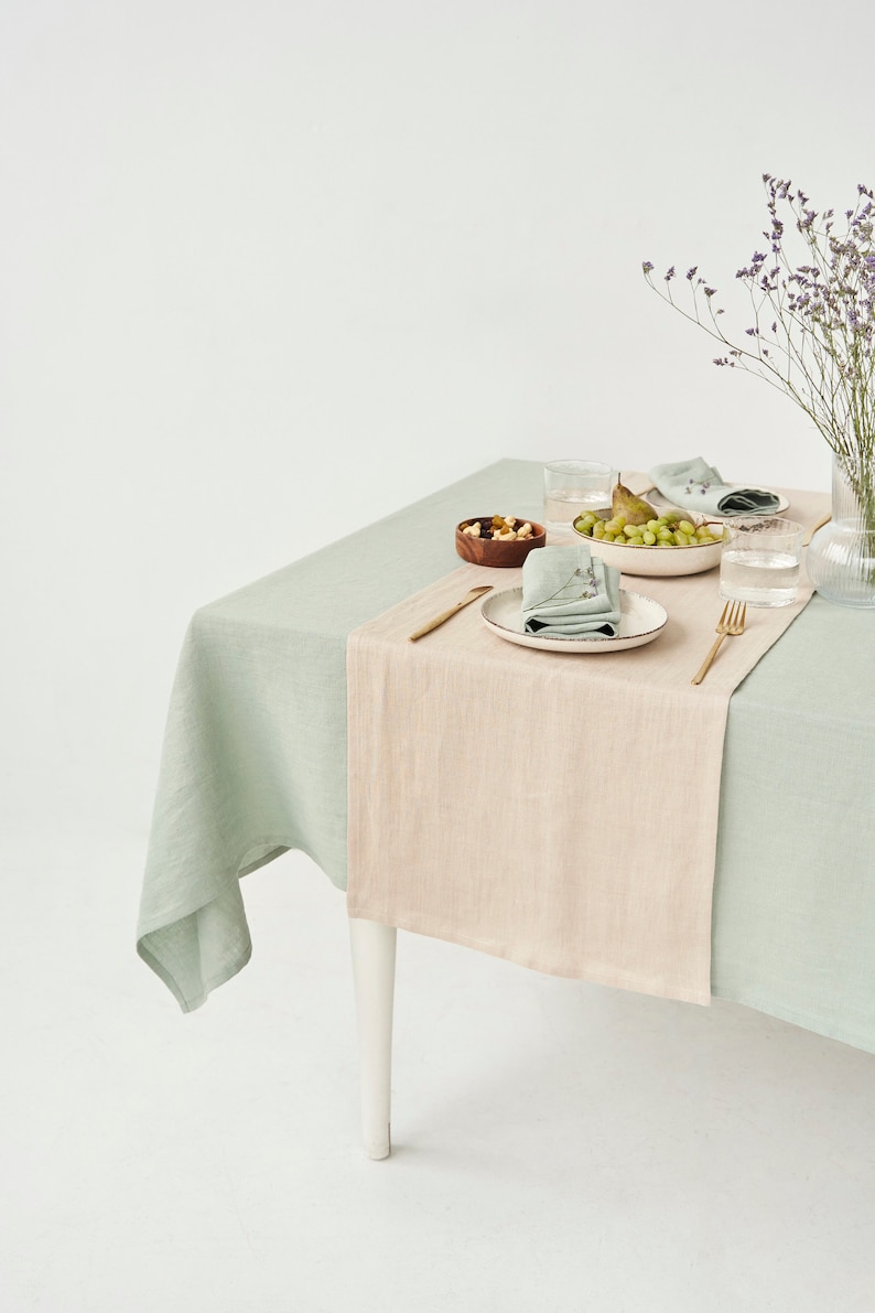 May include: A dining table set for a meal, featuring a light green tablecloth with a beige runner. The table is set with plates, napkins, glasses, and a bowl of fruit. A vase of purple flowers adds a decorative touch.