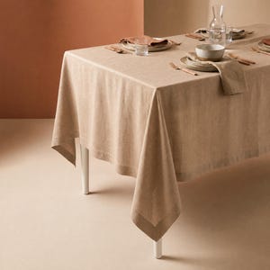 May include: A rectangular beige linen tablecloth draped over a table, set with white ceramic plates, bowls, clear glass tumblers, a glass carafe, and wooden cutlery. The table is positioned against a solid, muted orange-toned wall.