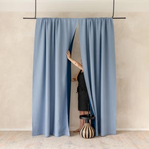 Puede incluir: Cortinas azul claro colgadas de una barra negra, con una persona parcialmente visible detrás. Las cortinas están ligeramente corridas, revelando un forro azul más oscuro. Una pequeña mesa decorativa con una base a rayas blancas y negras está en primer plano.