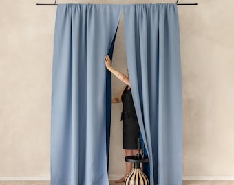 May include: A pair of floor-length, light blue curtains suspended from a black metal rod attached to the ceiling by chains. The curtains feature a solid, matte finish and hang in vertical folds, creating a clean, uniform appearance.