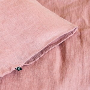 Puede incluir: Primer plano de una funda de almohada de lino rosa polvoriento con una peque&ntilde;a etiqueta negra. La tela tiene una textura sutil y un color suave y apagado.