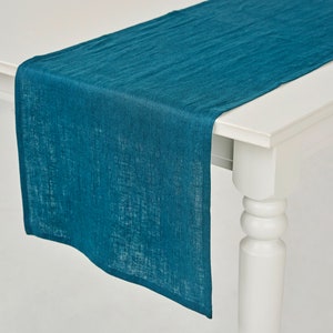 May include: Teal blue linen table runner. The runner is laying on a white table with a white table leg.