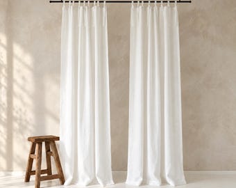 May include: Two floor-length white curtains hang from a black metal rod suspended by chains. The curtains feature tab tops and a simple, straight drape. A small, four-legged wooden stool sits on the floor to the left of the curtains.