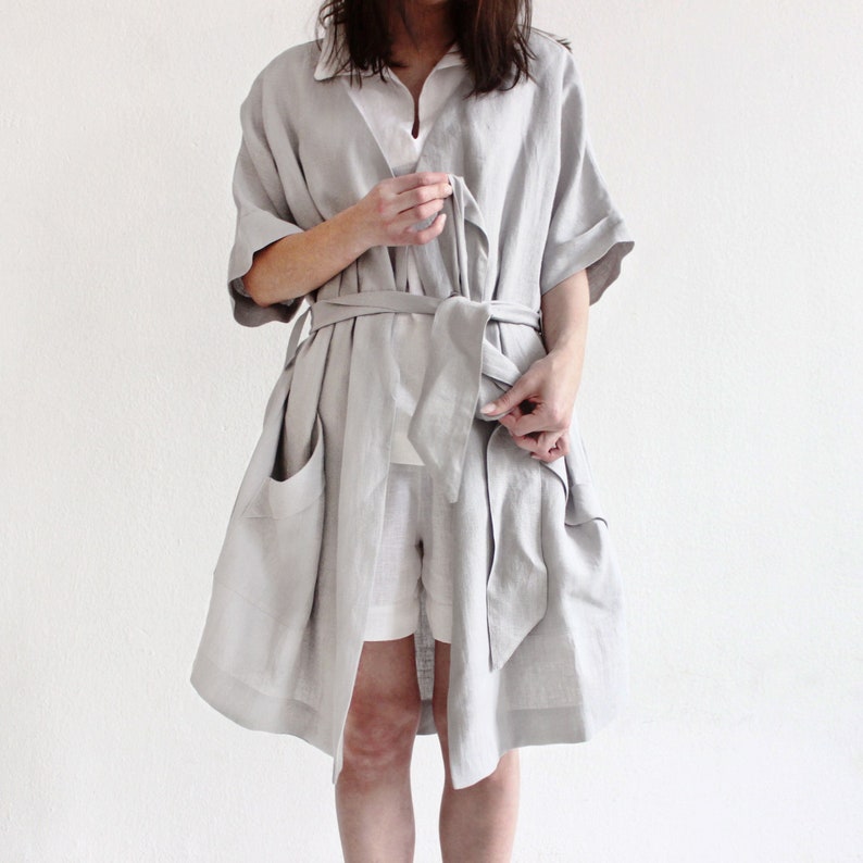 May include: A light grey linen robe with a self-tie belt and pockets. The robe is open at the front and the belt is tied at the waist.