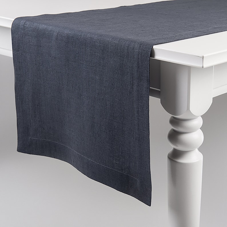 May include: A dark gray linen table runner with a subtle texture. The runner is laying on a white table with a turned leg.