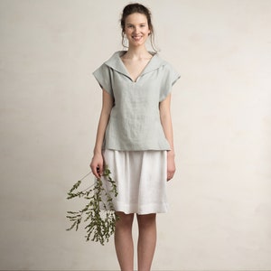 May include: A woman wearing a light blue linen top with a wide collar and a white linen skirt. She is holding a sprig of green foliage.