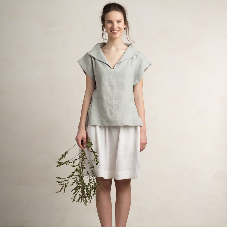 May include: A woman wearing a light blue linen top with a wide collar and a white linen skirt. She is holding a sprig of green foliage.